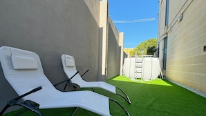 A patio area with two white lounge chairs and a small table, surrounded by artificial green turf and a beige wall. - Bayside (Marsaxlokk)
