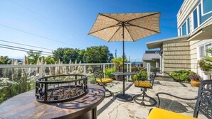 En udendørs terrasse med en rund bar, gule stole, en mønstret parasol og potteplanter.