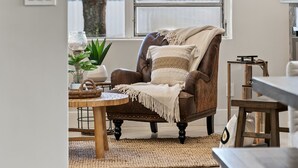 Un salon chaleureux avec un fauteuil en cuir, une table basse ronde en bois et une vue sur l’extérieur grâce à une grande fenêtre.