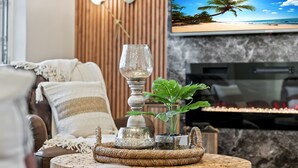 Un salon chaleureux avec un canapé en cuir marron, une table basse ronde en bois agrémentée d’objets décoratifs et un téléviseur diffusant une scène de plage.