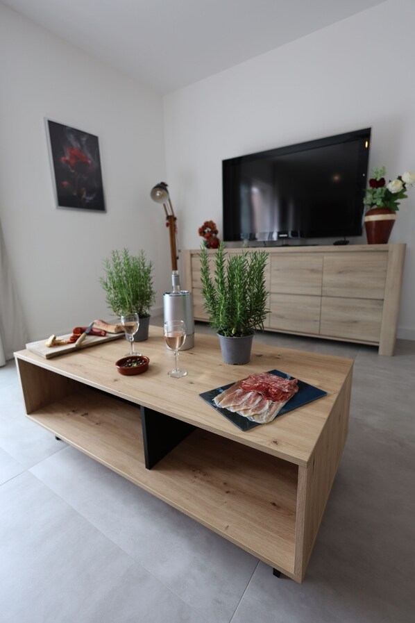 A modern living room with a wooden coffee table, a flat-screen TV, a potted plant, a wine glass, and a cutting board with food.
