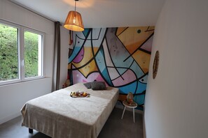 Ein Schlafzimmer mit einem Bett, einem Nachttisch, einer Lampe und einem farbenfrohen, abstrakten Wandbild.