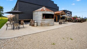A rustic outdoor seating area with tables and chairs, a metal barrel structure, and a building with a sign that reads 'THE VENUE.' - The Venue at Hermann – Room 7 Country Studio (Hermann)