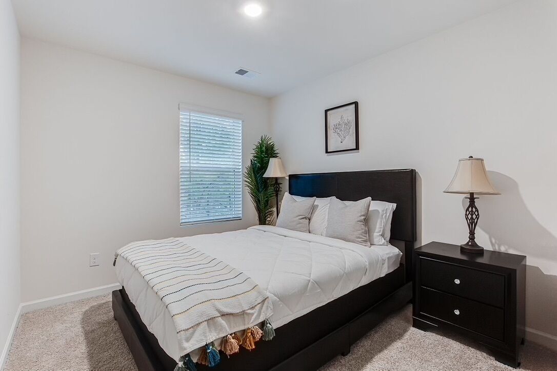 A bedroom with a bed, bedside tables, a window with blinds, a plant, and a framed picture on the wall.