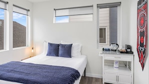 A bedroom with a bed, a nightstand, a desk with a kettle, and a window with blinds. - Dreamfyre's Ancient Dragon Horn Suite (Seattle)