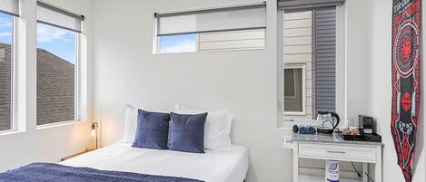 A bedroom with a bed, a nightstand, a desk with a kettle, and a window with blinds.