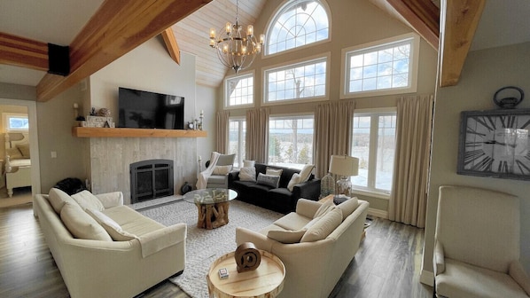 A spacious living room with a fireplace, large windows, and a chandelier.