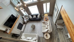 A spacious living area with a black sectional sofa, a beige sofa, and a wooden coffee table. There is a flat-screen TV mounted on the wall, and a chandelier hangs from the ceiling.