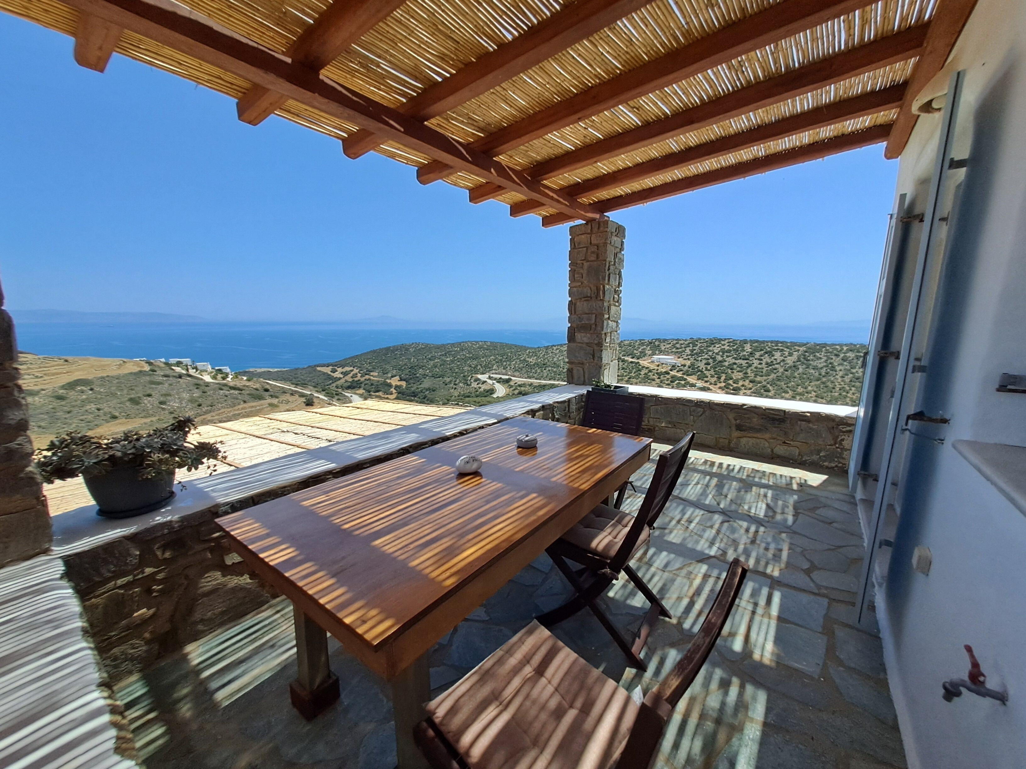Ein Holztisch mit Stühlen auf einer Steinterrasse mit Blick auf das Meer und die Hügel.