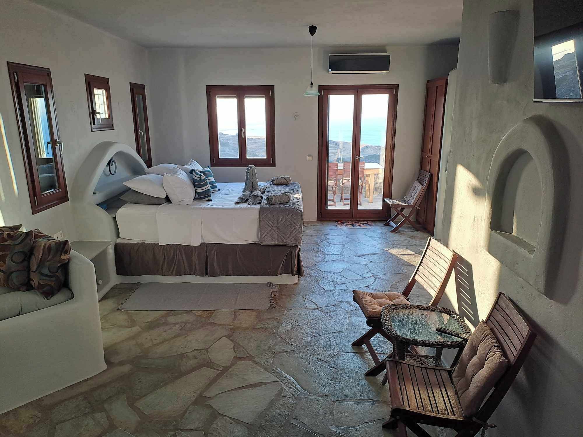 A bedroom with a bed, a sofa, a small table, and a chair. There are windows and a door.