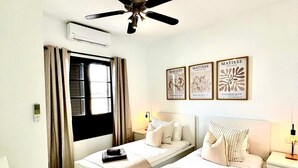 A hotel room with two beds, a ceiling fan, air conditioning, a window with curtains, bedside tables with lamps, and wall art. - Stunning 3 Bed 2 Bath Villa in Playa Blanca (Yaiza)