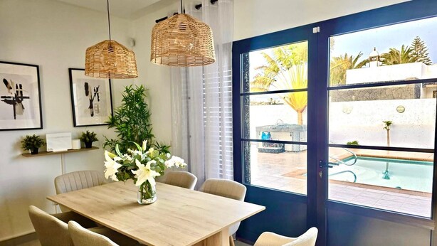 A dining area with a wooden table and chairs, a vase of flowers, and a view of a swimming pool through a glass door. - Stunning 3 Bed 2 Bath Villa in Playa Blanca (Yaiza)