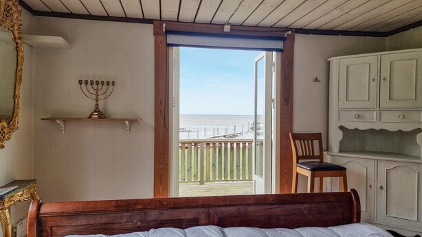 Een slaapkamer met een bed, een houten hoofdbord, een nachtkastje, een witte kast, een houten stoel, een menorah op een plank en een balkon met uitzicht op de zee.