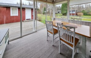 A covered patio with a wooden table and chairs, a red building in the background, and a view of a garden. - 3 bedroom pet friendly home in Källby (Källby)