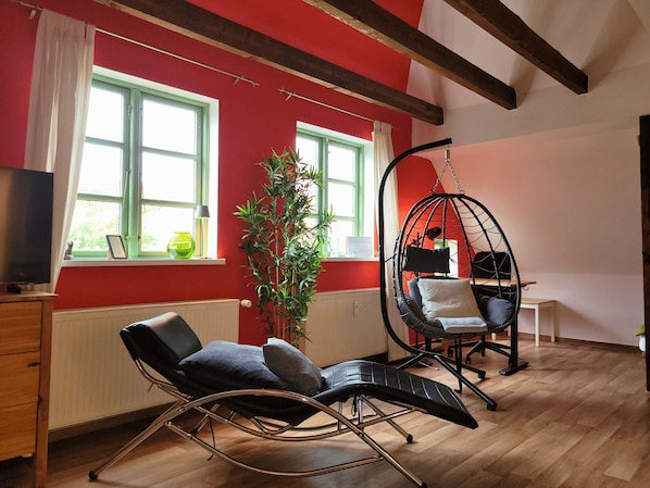 A room with a red wall, a black chair, a hanging chair, a wooden floor, and a window with a plant. - Marktstübchen: In the middle of the old town and wonderfully quiet (Waren (Müritz))