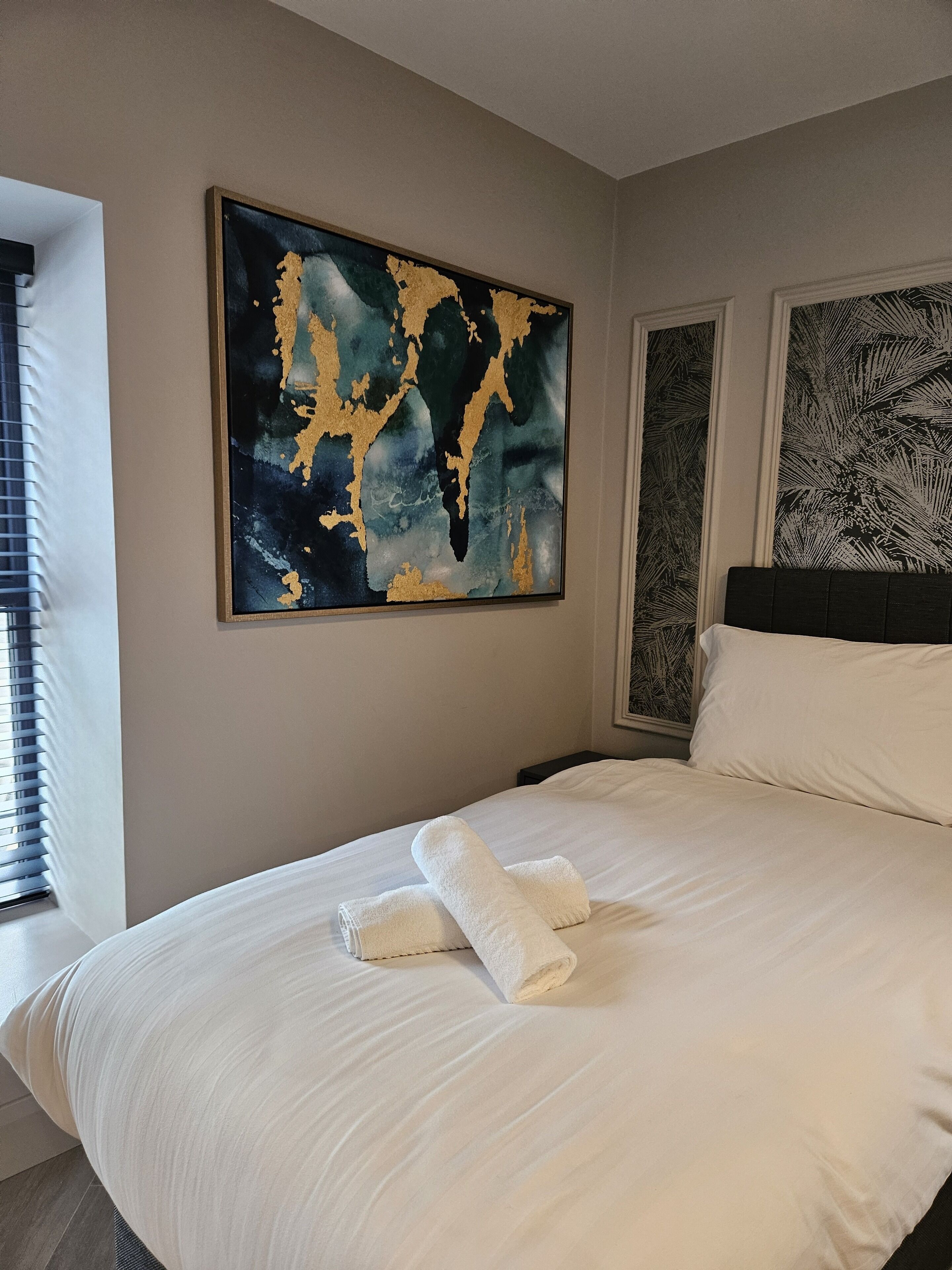 A hotel room with a large bed, white linens, a framed abstract painting, and a tropical leaf-patterned wall art.