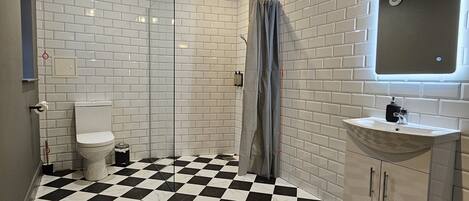 A bathroom with a checkered floor, white tiled walls, a toilet, a shower, and a sink with a mirror.