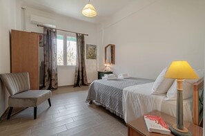 A hotel room with a bed, a chair, a bedside table with a lamp, a window with curtains, and a radiator. - Charming Central Corfu Apartment (Corfu)