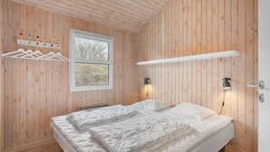Une pièce aux murs lambrés, comprenant un lit, une fenêtre, un porte-manteau et deux lampes fixées au mur.