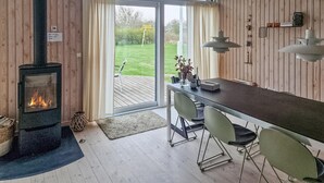Une pièce chaleureuse avec un poêle à bois, une table à manger avec des chaises et une vue sur un jardin, visible à travers de grandes portes vitrées.