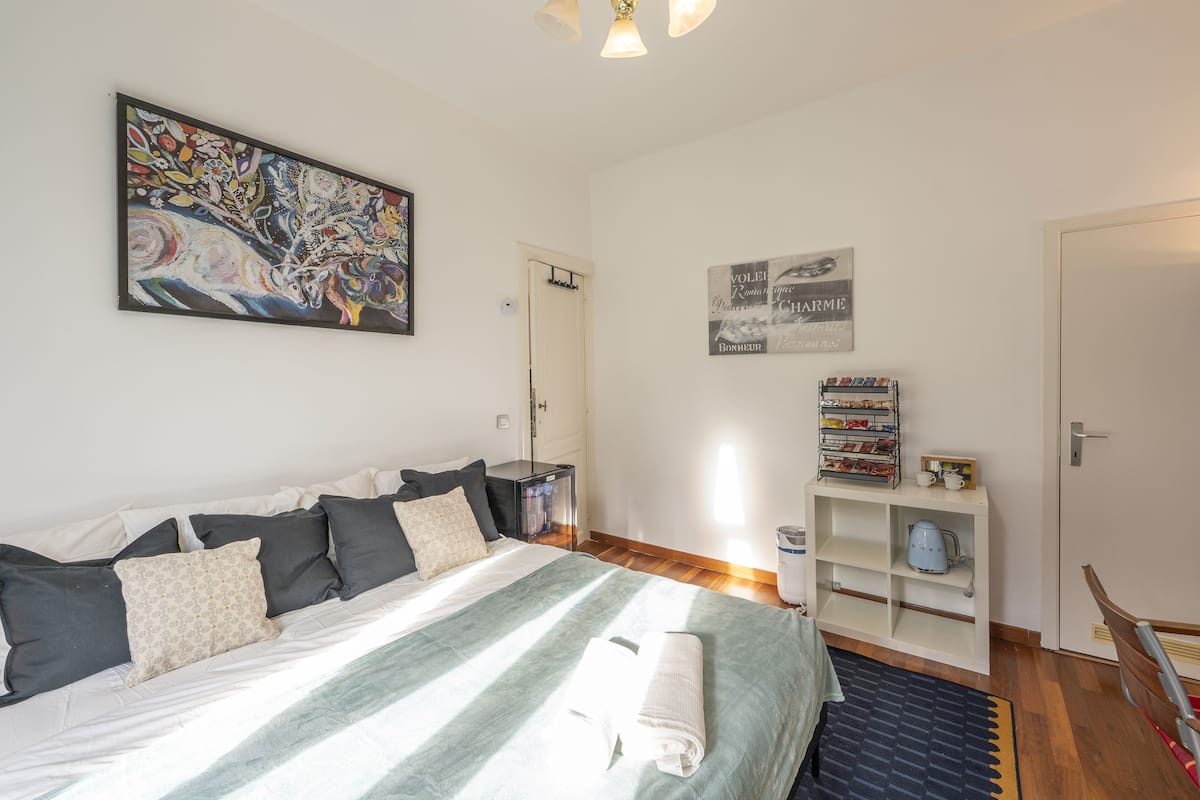 A neatly arranged bedroom with a bed, a painting, a shelf with toiletries, and a door.