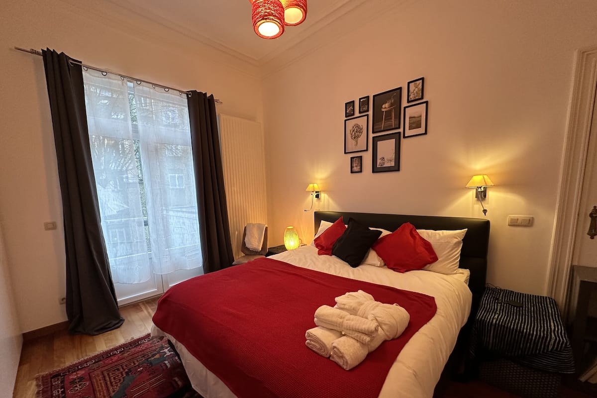 A hotel room with a large bed, two bedside lamps, a window with curtains, and a wall with framed pictures.