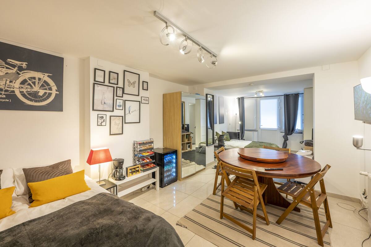 A modern hotel room with a bed, a dining table with chairs, a refrigerator, a coffee machine, and a wall with framed pictures.