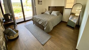 A bedroom with a large bed, a wooden floor, a small bedside table, a nightstand, a mirror, and a view of the outdoors through a sliding door. - Estancia Cataviña Valle de Guadalupe (Valle de Guadalupe)