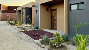 A modern building with a minimalist design, featuring a concrete facade, a wooden door, and a small garden area with rocks and plants. - Estancia Cataviña Valle de Guadalupe (Valle de Guadalupe)