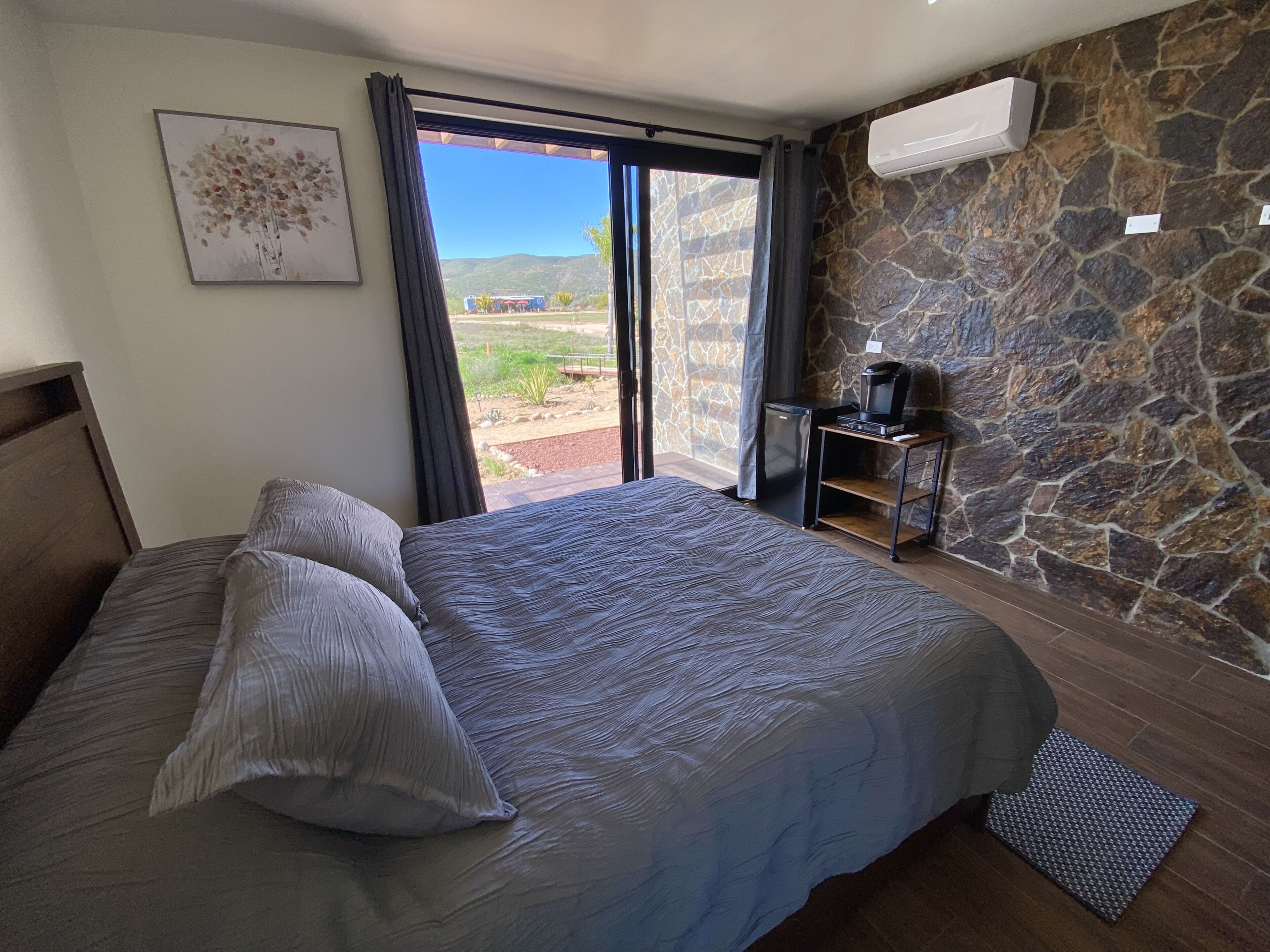 Ein Schlafzimmer mit Bett, einem kleinen Tisch mit Kaffeemaschine, einer Steinwand und Blick ins Freie.