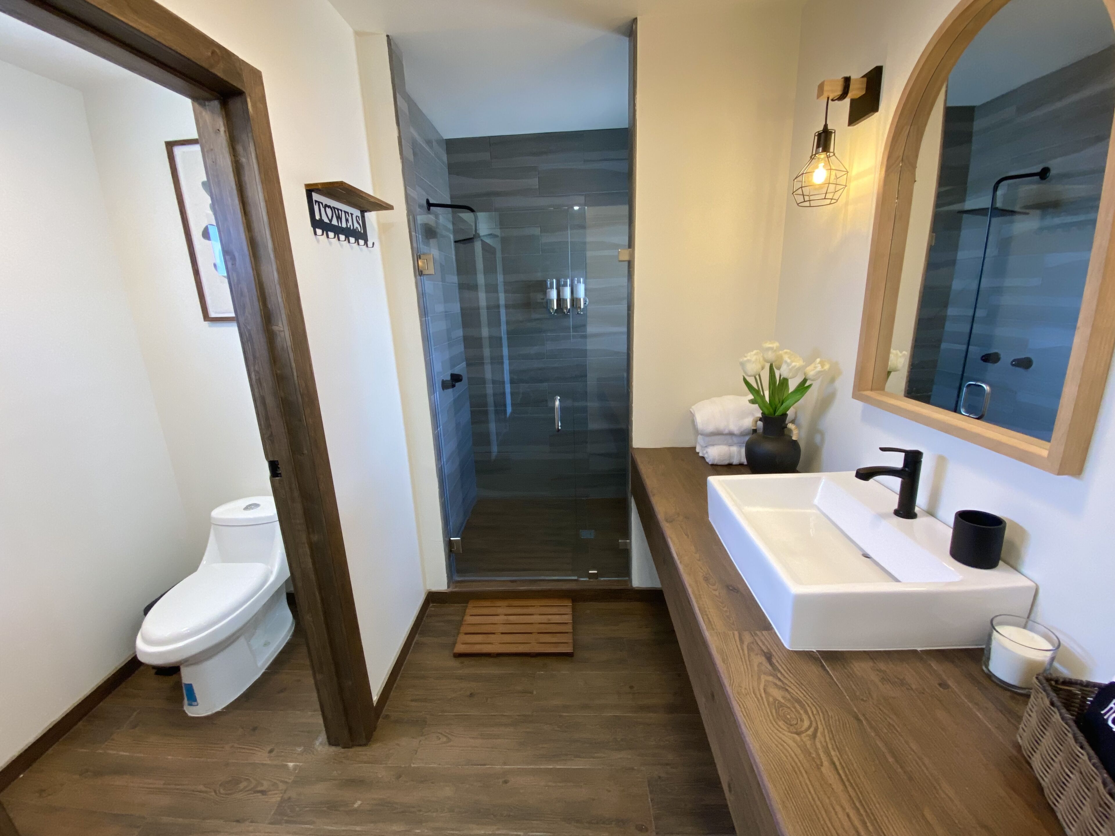 A modern bathroom with a white toilet, a glass shower enclosure, a wooden vanity with a sink, and a mirror.