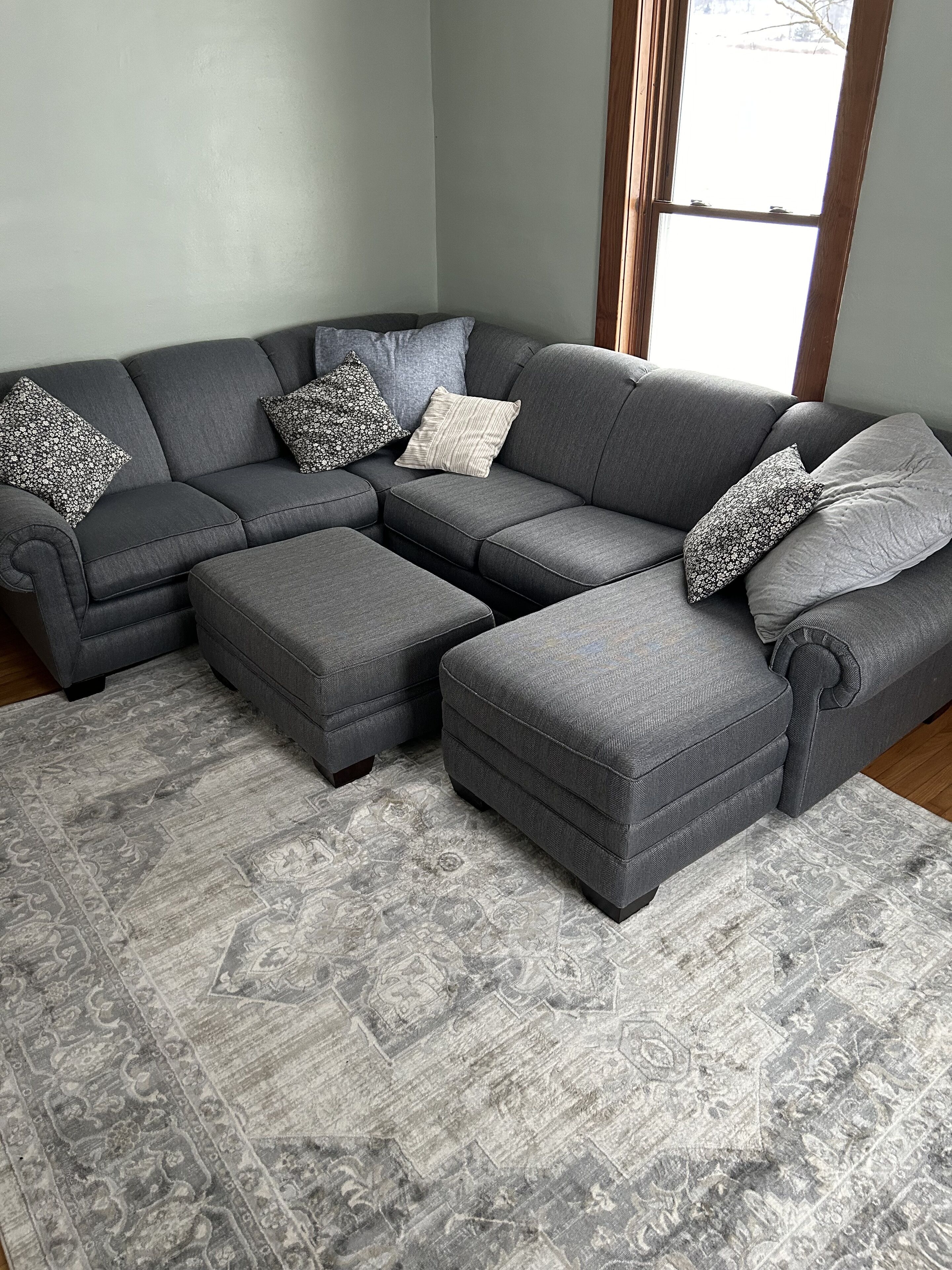 A grey sectional sofa with matching ottomans and patterned throw pillows, set on a grey patterned rug in a room with a large window.