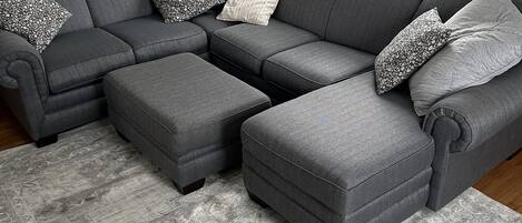 A grey sectional sofa with matching ottomans and patterned throw pillows, set on a grey patterned rug in a room with a large window.