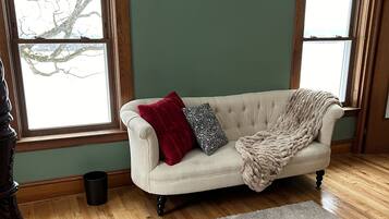A room with a beige sofa, a red and black pillow, a beige blanket, and a plaid rug. There are two windows with wooden frames and a view of bare tree branches.