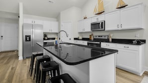 A modern kitchen with white cabinetry, a black countertop island, stainless steel appliances, and a wooden floor. - Modern NC Home: Dock, Karaoke, Games, Fire Pit (Oriental)