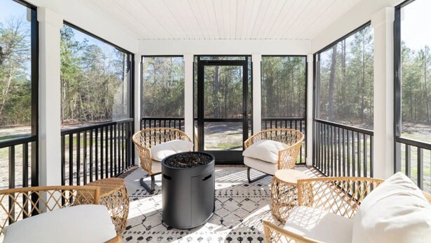 A covered outdoor area with wicker furniture, a black trash can, and large windows offering a forest view. - Modern NC Home: Dock, Karaoke, Games, Fire Pit (Oriental)