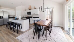 A modern kitchen with a central island, stainless steel appliances, and a dining area with a wooden table and chairs. - Modern NC Home: Dock, Karaoke, Games, Fire Pit (Oriental)