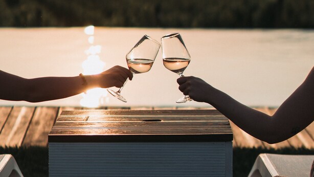 Two people toasting with wine glasses at a wooden table with a view of a body of water and a bridge in the background. - METHE Resort (Lizzano)