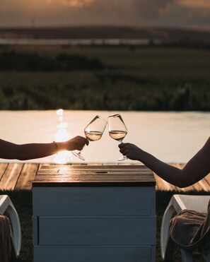 Two people toasting with wine glasses at a wooden table with a view of a body of water and a bridge in the background.