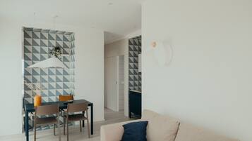 A modern living room with a beige sofa, a dining area with a table and chairs, and a kitchen area visible through an open door.