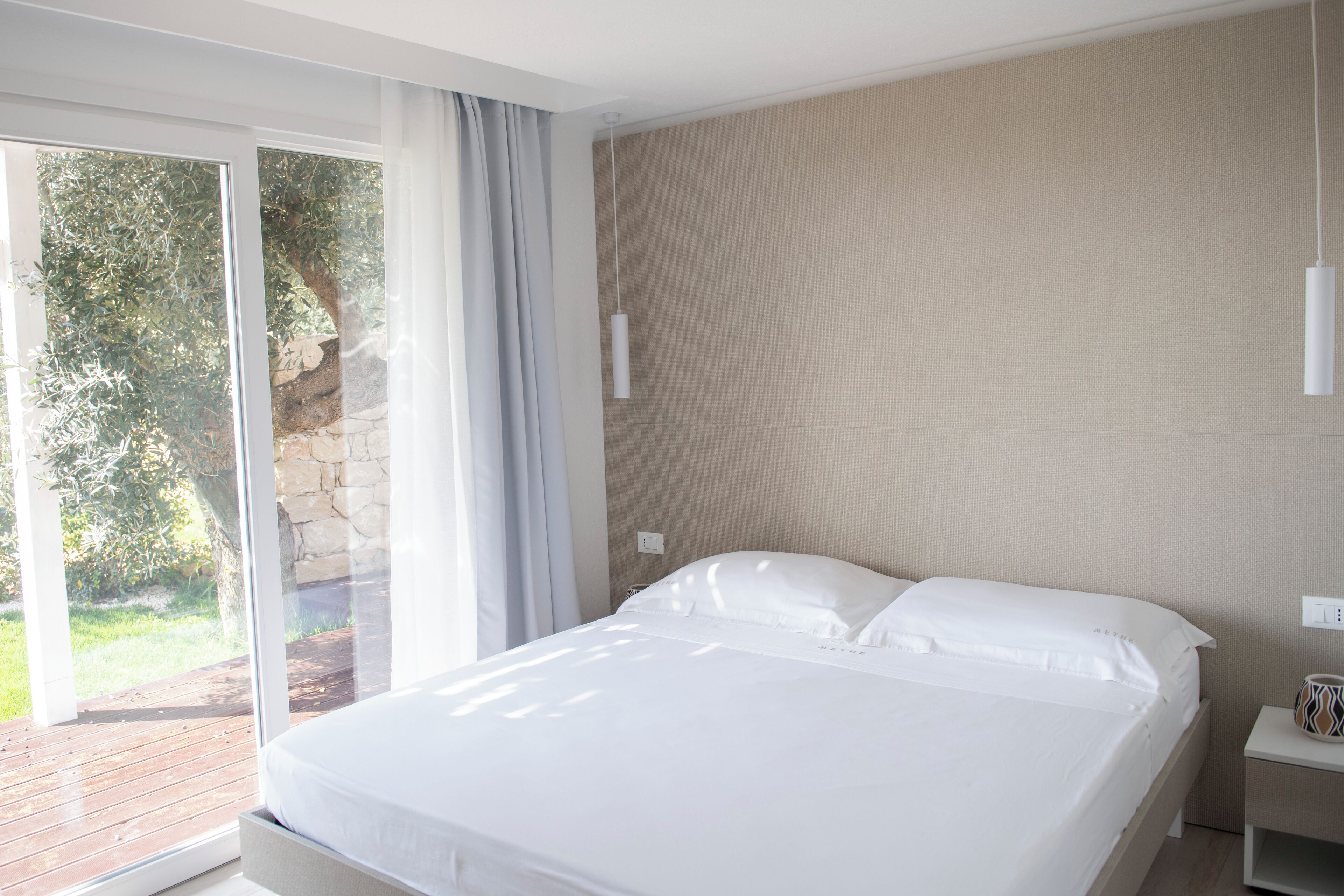 A modern hotel room with a large bed, a nightstand, and a view of a garden through a sliding glass door.