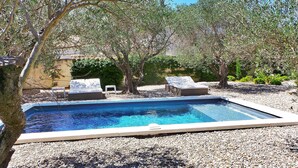 Piscina com duas espreguiçadeiras e uma pequena mesa, rodeada por oliveiras e pedrinhas.