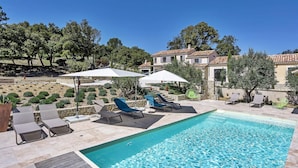 A pool area with lounge chairs and umbrellas, surrounded by trees and a building with a tiled roof. - Lovely home in Roussas (Roussas)