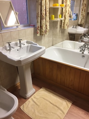 A bathroom with a sink, a bidet, a bathtub, wooden paneling, a mirror, and a window with floral curtains. - Lowri Cottage Hotel (Birmingham)