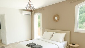 Une chambre avec un grand lit, un luminaire suspendu, un miroir rond et une fenêtre donnant sur un paysage verdoyant.