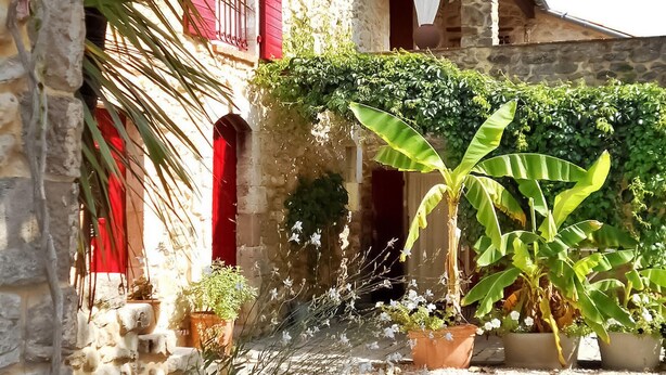 A stone building with red shutters, a small water feature, and various potted plants.