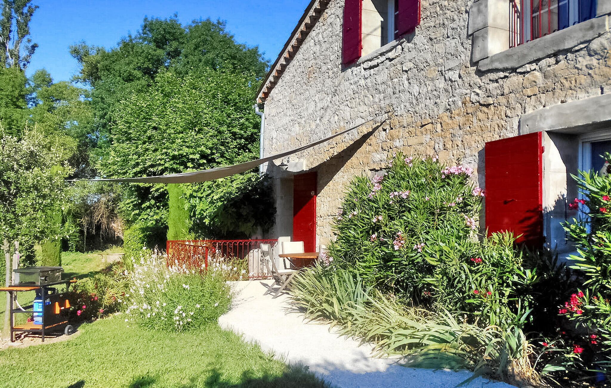 A stone house with a red door and window shutters, surrounded by lush greenery and flowering plants.