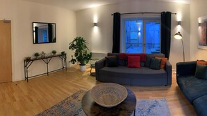 Een moderne woonkamer met een grijze bank, een ronde salontafel, een grote potplant en uitzicht buiten door een raam met zwarte gordijnen.