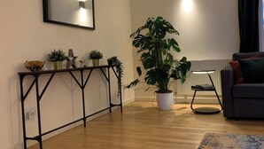 Een woonkamer met een houten vloer, een console tafel met planten, een grijze bank en een grote kamerplant.
