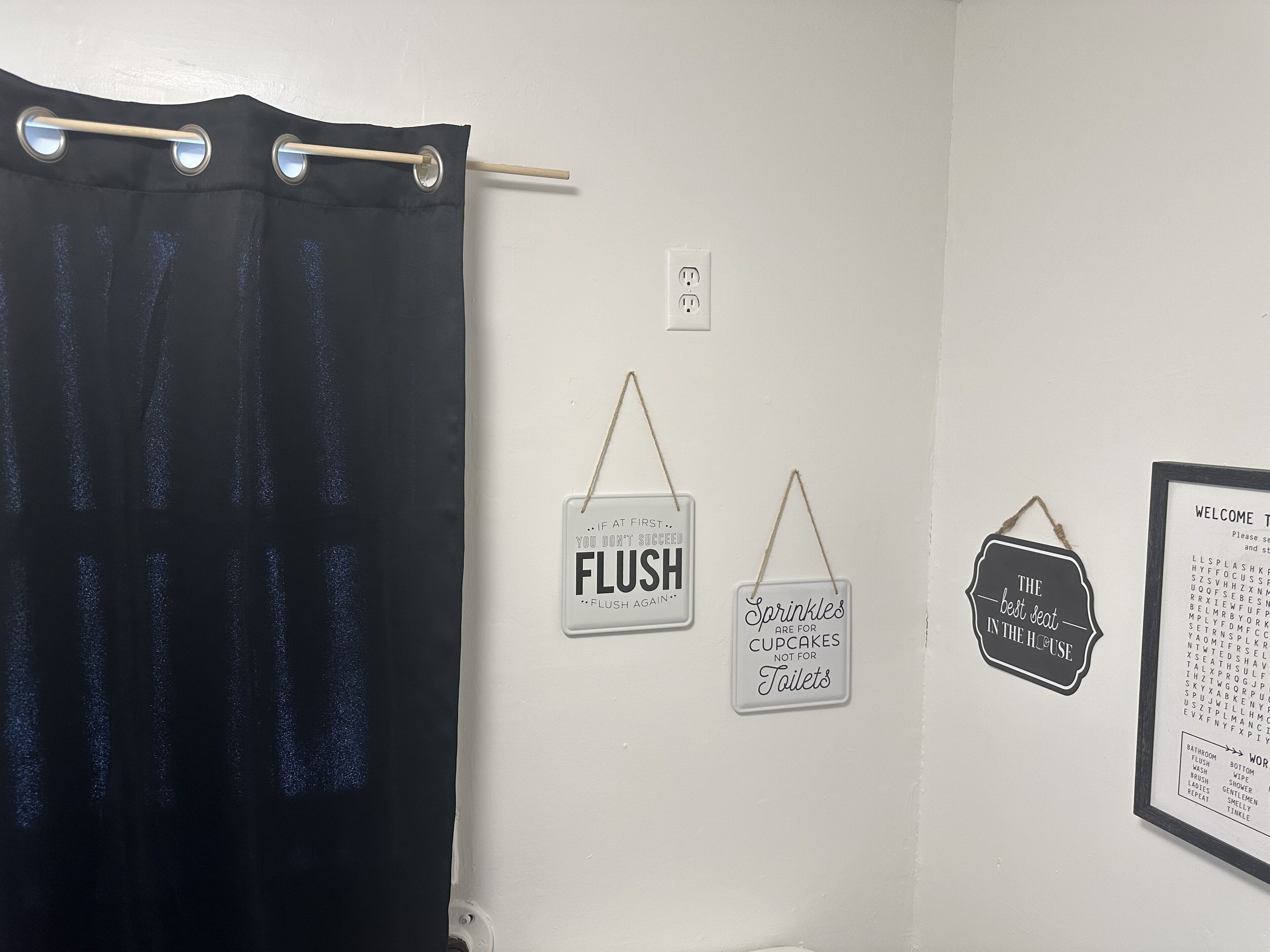 Un bany amb cortina de bany negra, un rètol fixat a la paret que diu "FLUSH" i un cartell de benvinguda emmarcat amb una llista de serveis del bany.